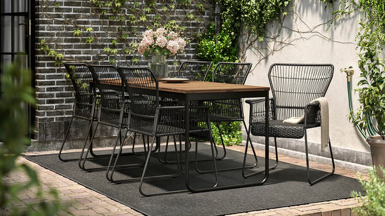A vase full of pink flowers sits on a wooden table surrounded by black chairs in a plant-filled courtyard