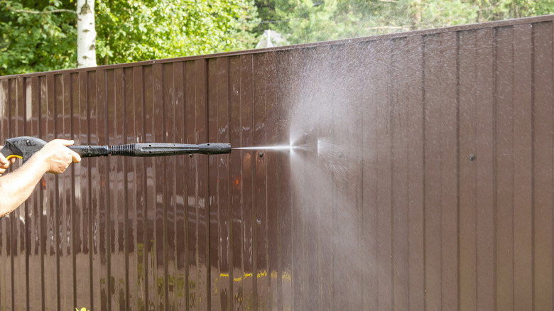 Hands holding a pressure washer wand and cleaning a vinyl fence or wall