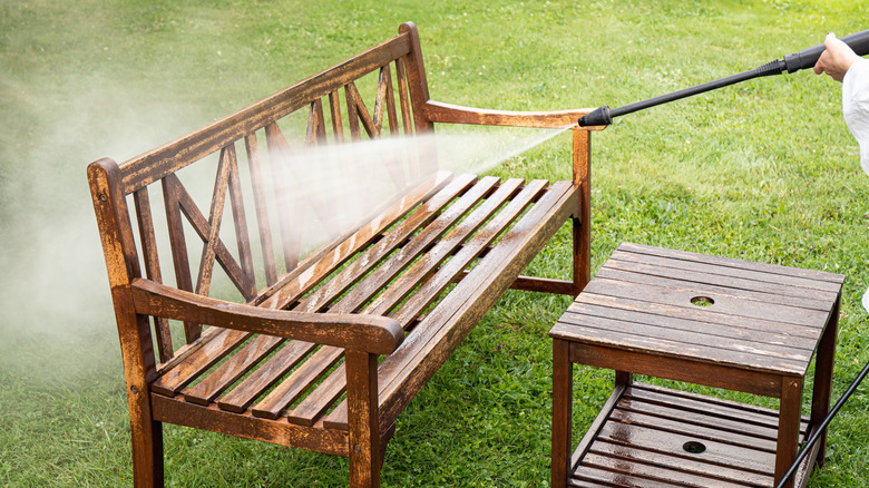 Hand holding a pressure washer wand to clean a wood bench on green grass