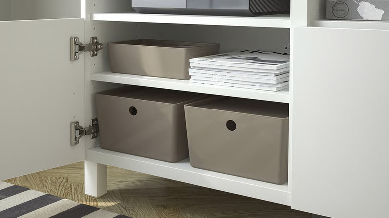 A white cabinet door is opened to reveal a stack of magazines and three beige IKEA storage boxes