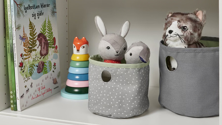 Two gray fabric IKEA containers filled with stuffed animals sit next to children's books and toys
