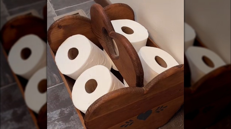 Four toilet paper rolls tucked into dark wood magazine holder on bathroom tile