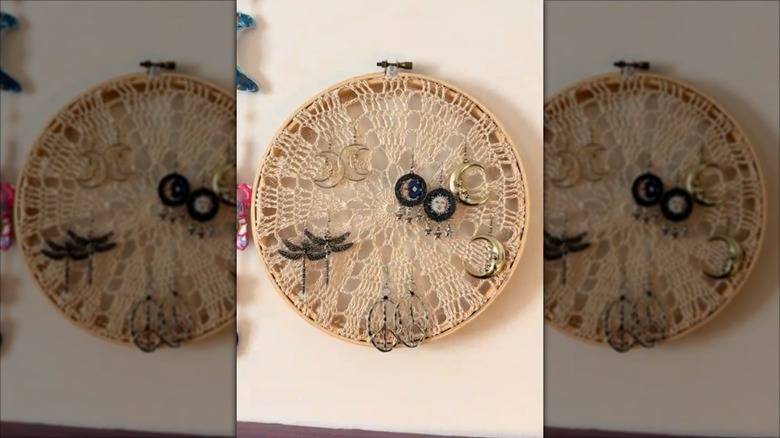 A doily in an embroidery hoop hung on the wall with earrings hanging from it.