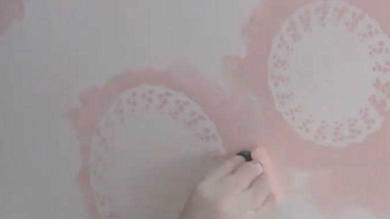 A wall with doilies glued to it while a person paints the wall around them pink.