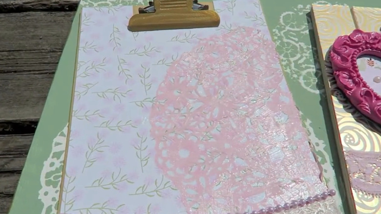 A clipboard decorated with floral paper, a doily, and beads.