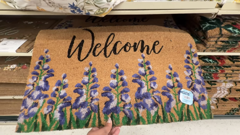 A person pulls a floral coir doormat that reads 'Welcome' off a shelf at Hobby Lobby