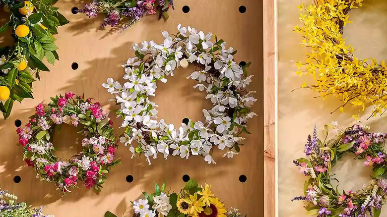 A faux white dogwood wreath hangs on a wooden wall surrounded by other colorful floral wreaths