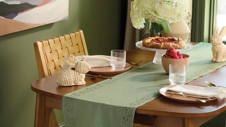 A green eyelet table runner sits on a wooden dining table decorated with flowers and woven bunny statues