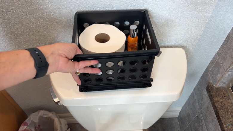 Woman stocking toilet paper and room spray in milk crate behind toilet