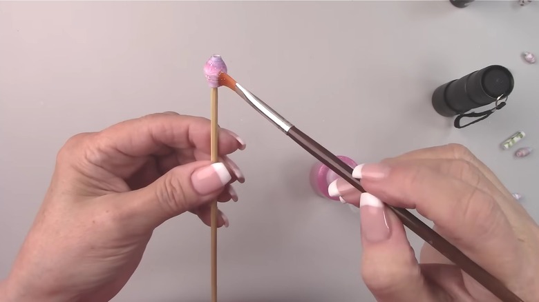 Woman painting paper bead on skewer over white table with other handcrafted beads