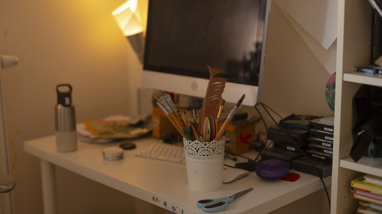 A cluttered home office desk with scissors, papers, discs, and paint brushes taking up space