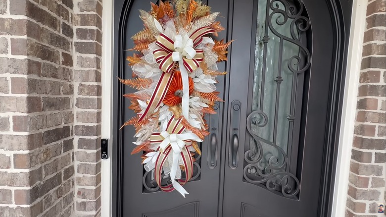 Fall-themed teardrop swag on arched ornate front door beside brick wall