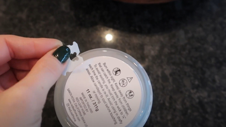A hand using a white bread tag to scrap the label off of a candle.