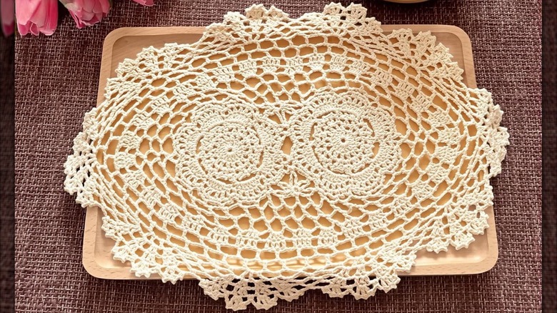 Oval doily laying on wooden decorative tray on woven placemat beside pink flowers