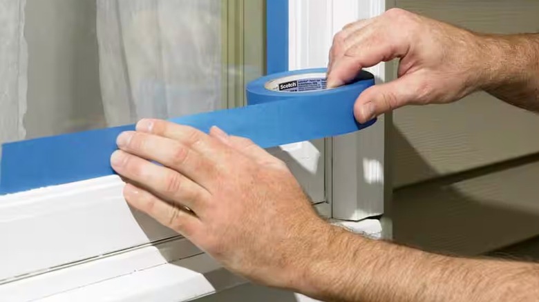 A person's hands are shown applying blue painter's tape to a window frame