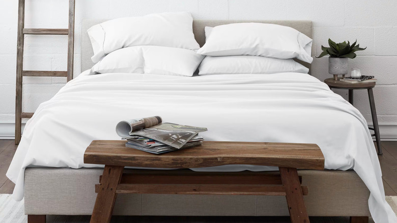 A wooden bench is in front of a modern bed with white sheets