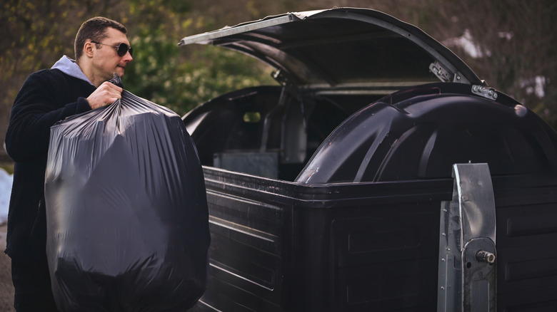 A man heaves a large black trash bag into a large lidded dumpster