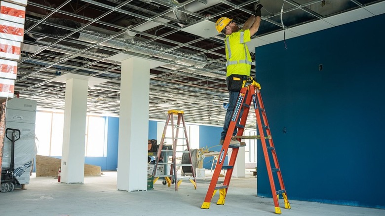 A man stands on an orange Werner fiberglass step ladder while working in a ceiling