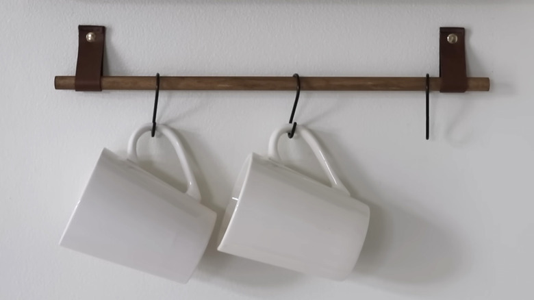 Two white coffee cups hang from hooks on a simple leather and wooden dowel wall display
