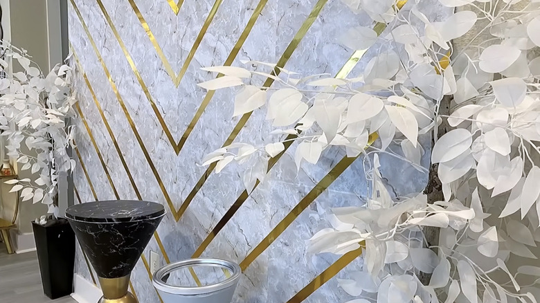 Potted artificial plants sit in front of a marble and gold chevron wallpaper-covered accent wall
