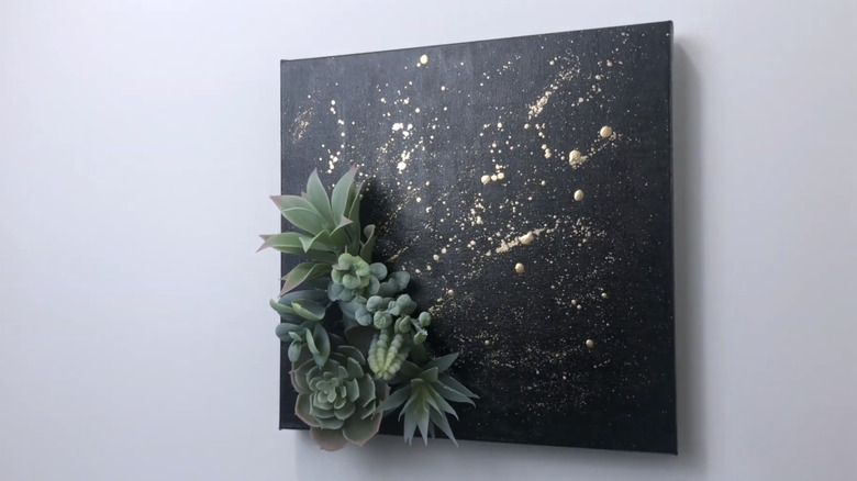 Small succulents are tucked into the bottom corner of a black canvas splattered with gold paint