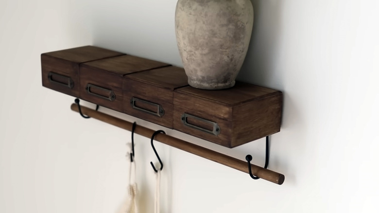 A clay vase sits on a simple shelf made of stained wooden drawers and a thin dowel