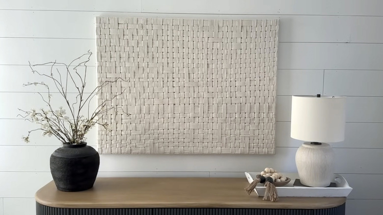 A large piece of woven wall art hangs over a console table between a black vase and white lamp
