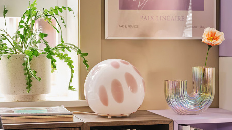 A soft pink and white FADO table lamp from IKEA sits on a shelf between a potted plant and a bud vase