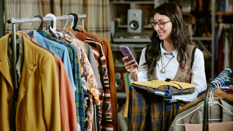 Smiling, young woman thrifting many kinds of clothes while browsing her phone for ideas
