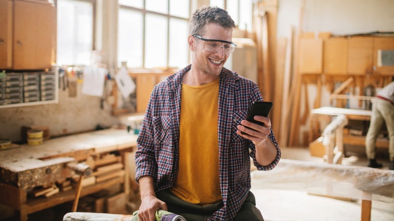 A man smiling in his workshop while shopping for new tools on his phone