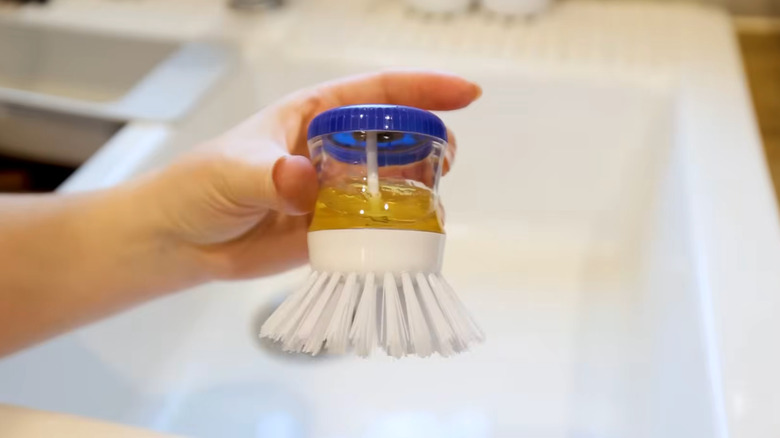 A person holds a Dollar Tree Scrub Buddies Soap Dispensing Brush over a white sink