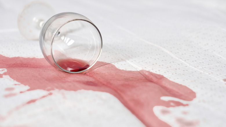 Wine glass overturned with red wine spilled on white fabric.