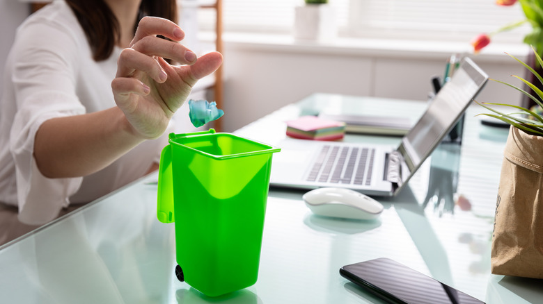 A person throwing trash into small, green trash bin in a home office
