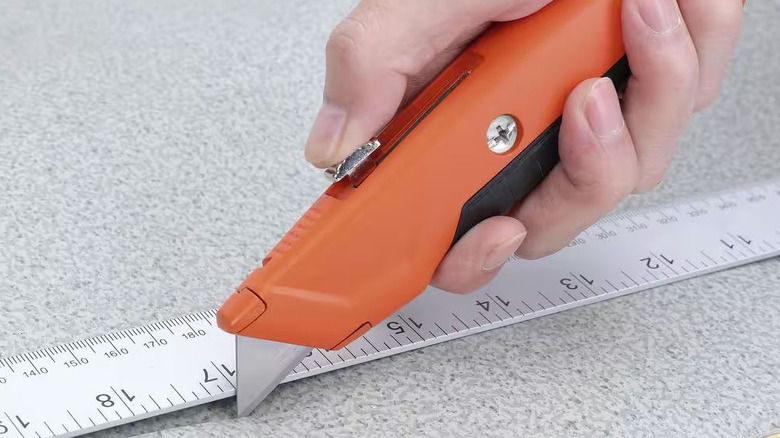 A person uses an orange retractable utility knife against a metal ruler to cut gray material