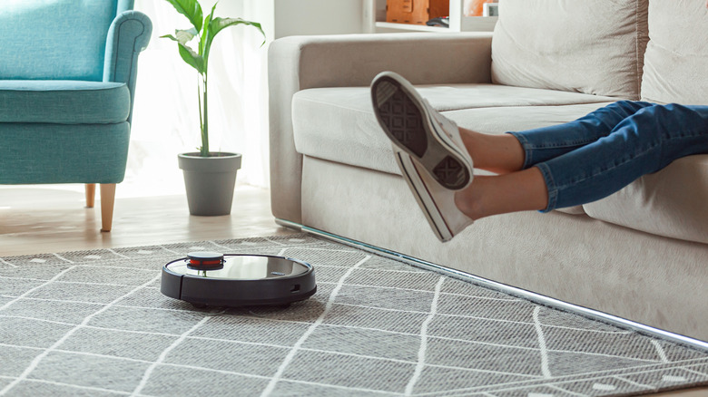 A person sits on a couch and lifts their feet as a robot vacuum cleans the floor beneath