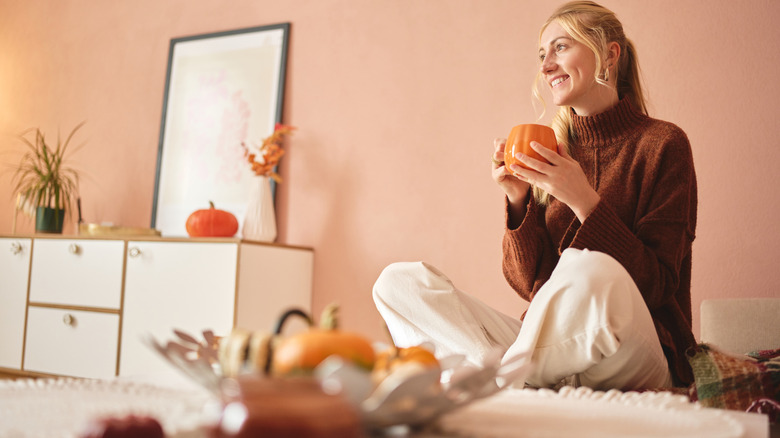 A smiling woman holds a pumpkin mug while thinking in a fall-themed room