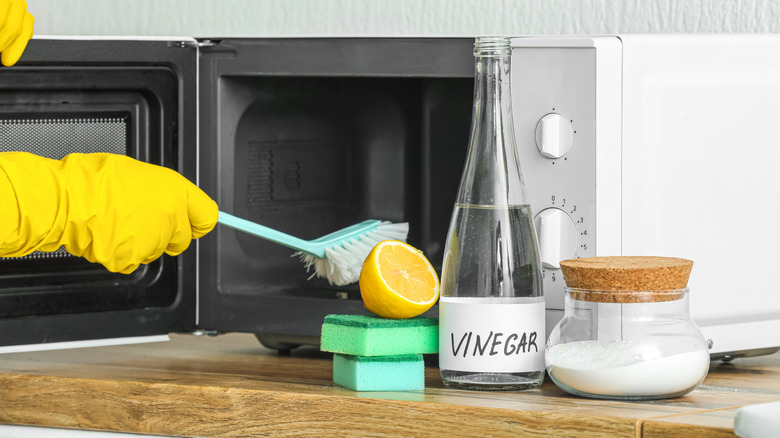 A gloved hand scrubbing the inside of a microwave with sponges, a lemon, a bottle of vinegar, and baking soda sitting nearby.