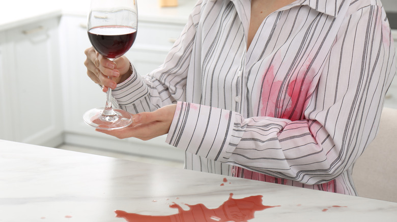 Woman holding a glass of red wine in the kitchen with spills on the counter and her shirt.
