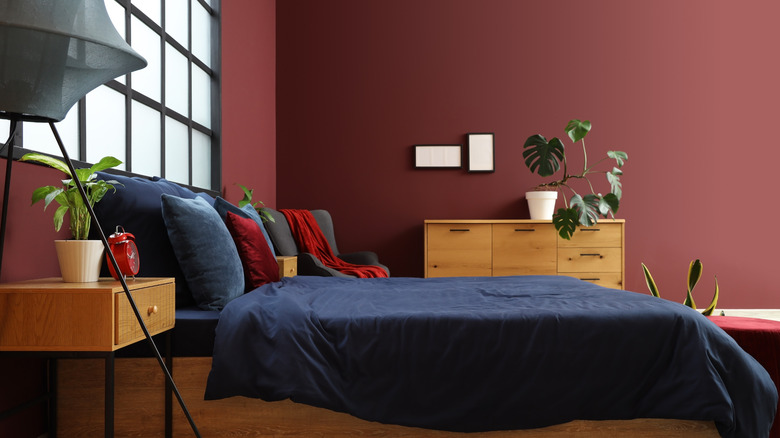 Burgundy walls in a bedroom with a wood dresser, black-framed window, and bed covered in navy linens