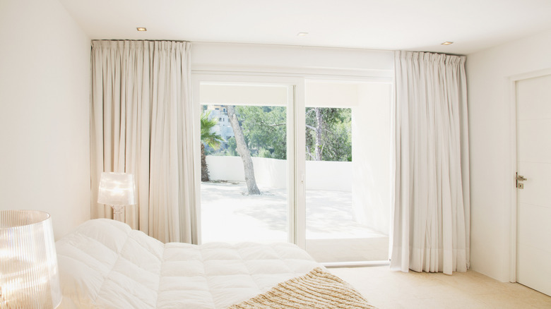 A minimalist white and light brown bedroom with single white curtains pulled open