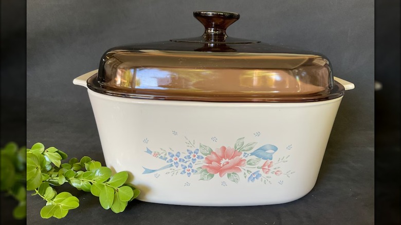 Vintage CorningWare Symphony casserole dish with Pyrex tinted glass top on gray backdrop with artificial foliage