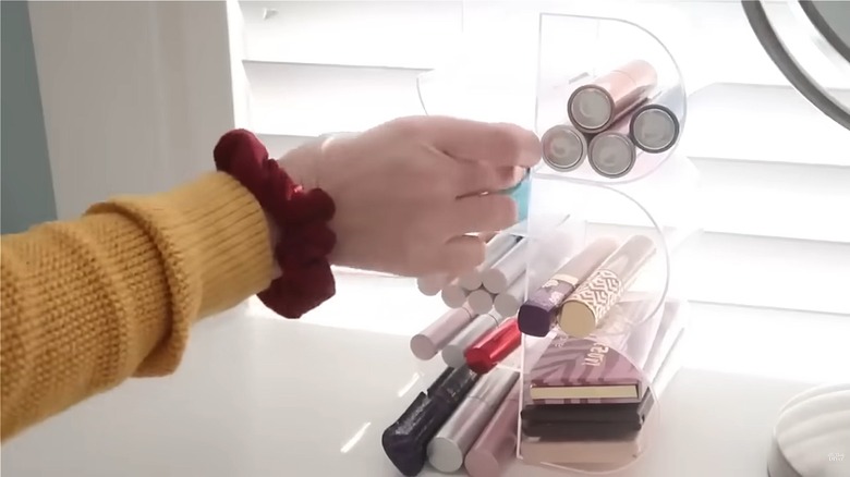 Woman tucking various makeup products into clear compartments of DIY Dollar Tree organizer in front of window on white surface