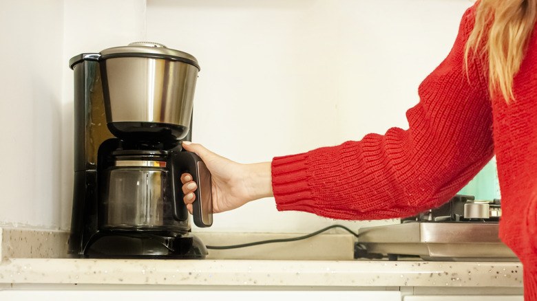 A person in a red sweater holds onto a coffeemaker on a plain kitchen counter.