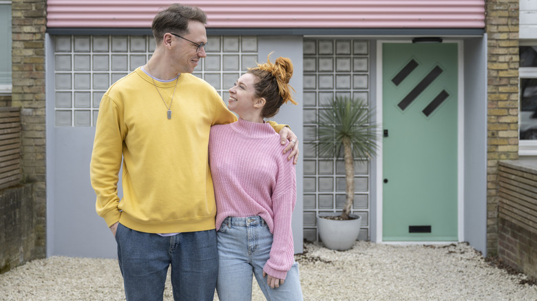 A couple stands outside a colorful house with a dated gravel driveway