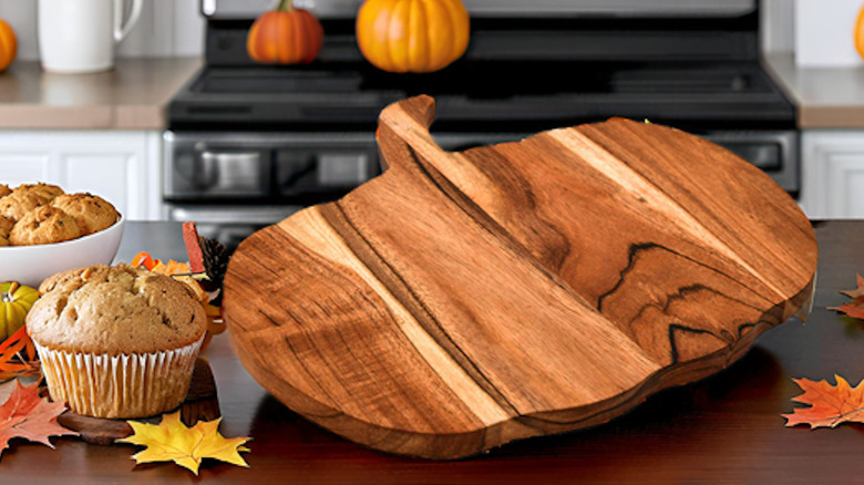 A pumpkin-shaped wooden cutting board on a kitchen island with fall accessories