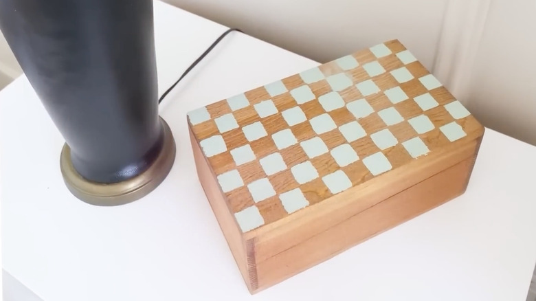 A wooden recipe box with green checkered paint on a white nightstand next to a black lamp