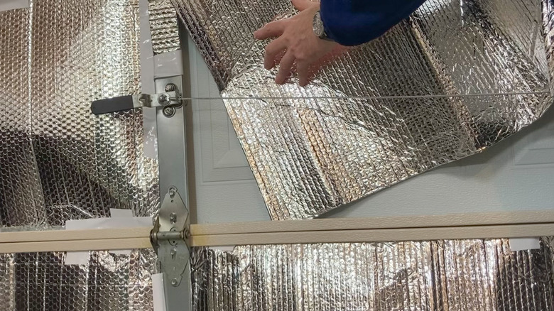 photo of a whiter person's arm wearing a watch and a blue sweater applying silver metallic insulation sheets against a garage door