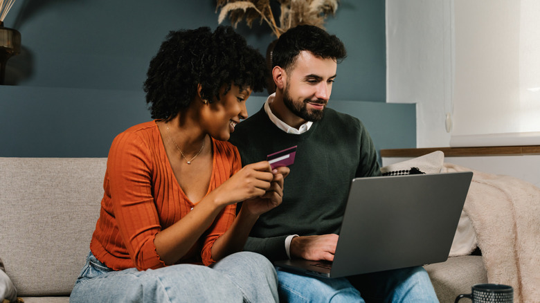A couple smiles as they sit on a sofa and shop online via a laptop