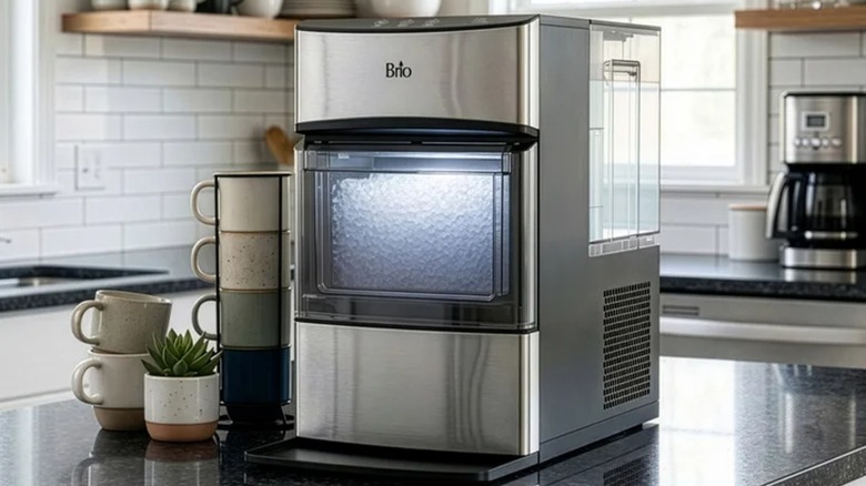 A stainless steel Brio nugget ice maker sits on a kitchen countertop next to a stack of coffee mugs