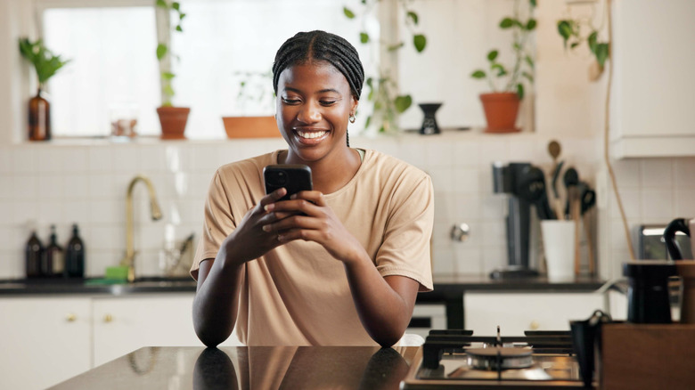 A woman scrolls on her phone in her kitchen while smiling